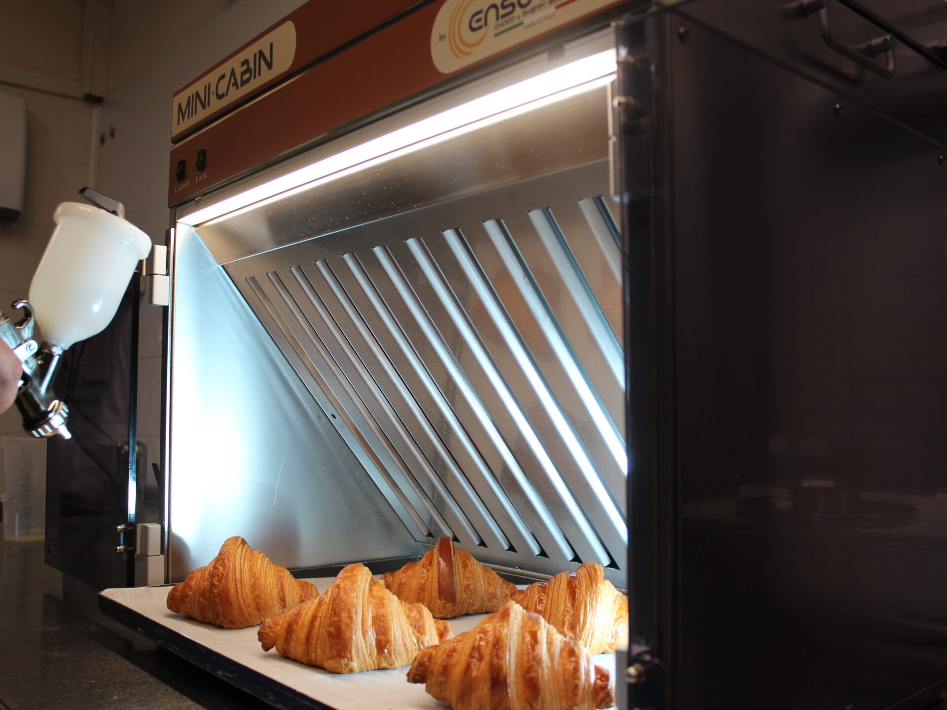 Mini Cabin Ensotech con croissant in fase di decorazione a spruzzo: cabina di aspirazione compatta per pasticceria e cioccolato.